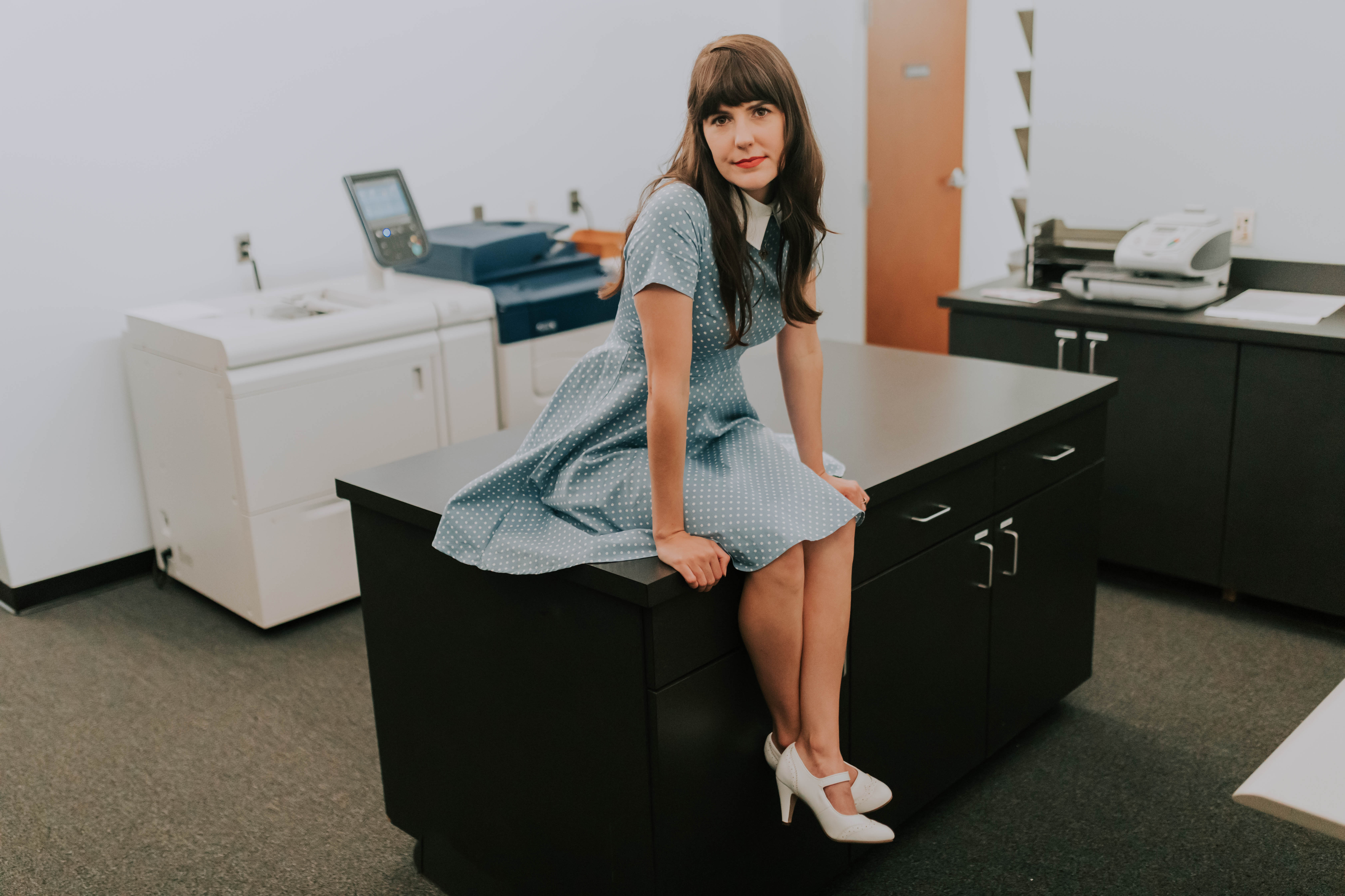 KellyMarie, wearing a dress, sits inside a office