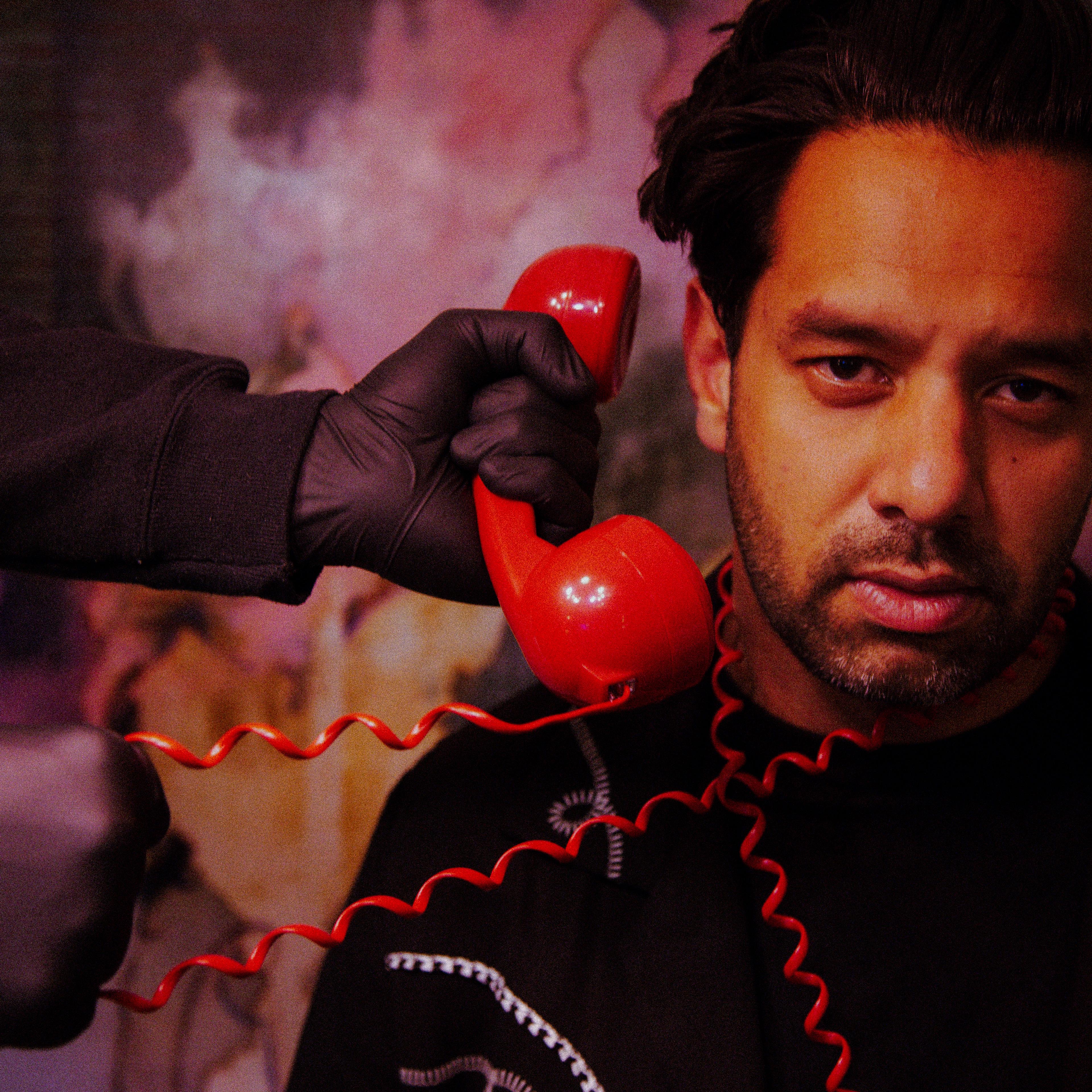A man with a red phone cord wrapped around him and another person with a black glove holding the phone to his ear