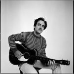 Holding an acoustic guitar and wearing a striped shirt, Andrew Combs faces the camera
