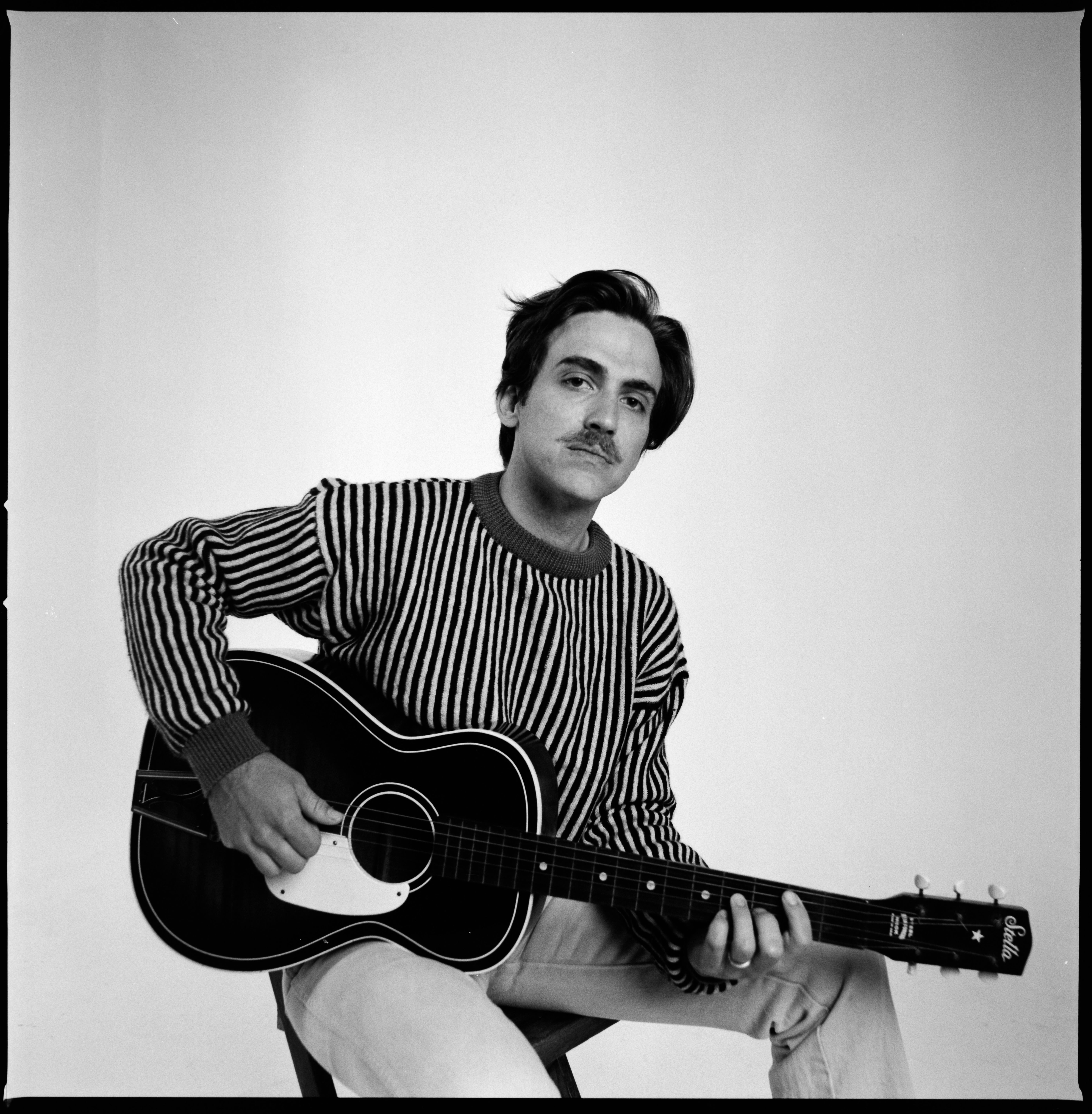 Holding an acoustic guitar and wearing a striped shirt, Andrew Combs faces the camera