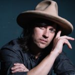 With a fedora on his head, Rhett Miller, in a dark shirt, looks at the camera