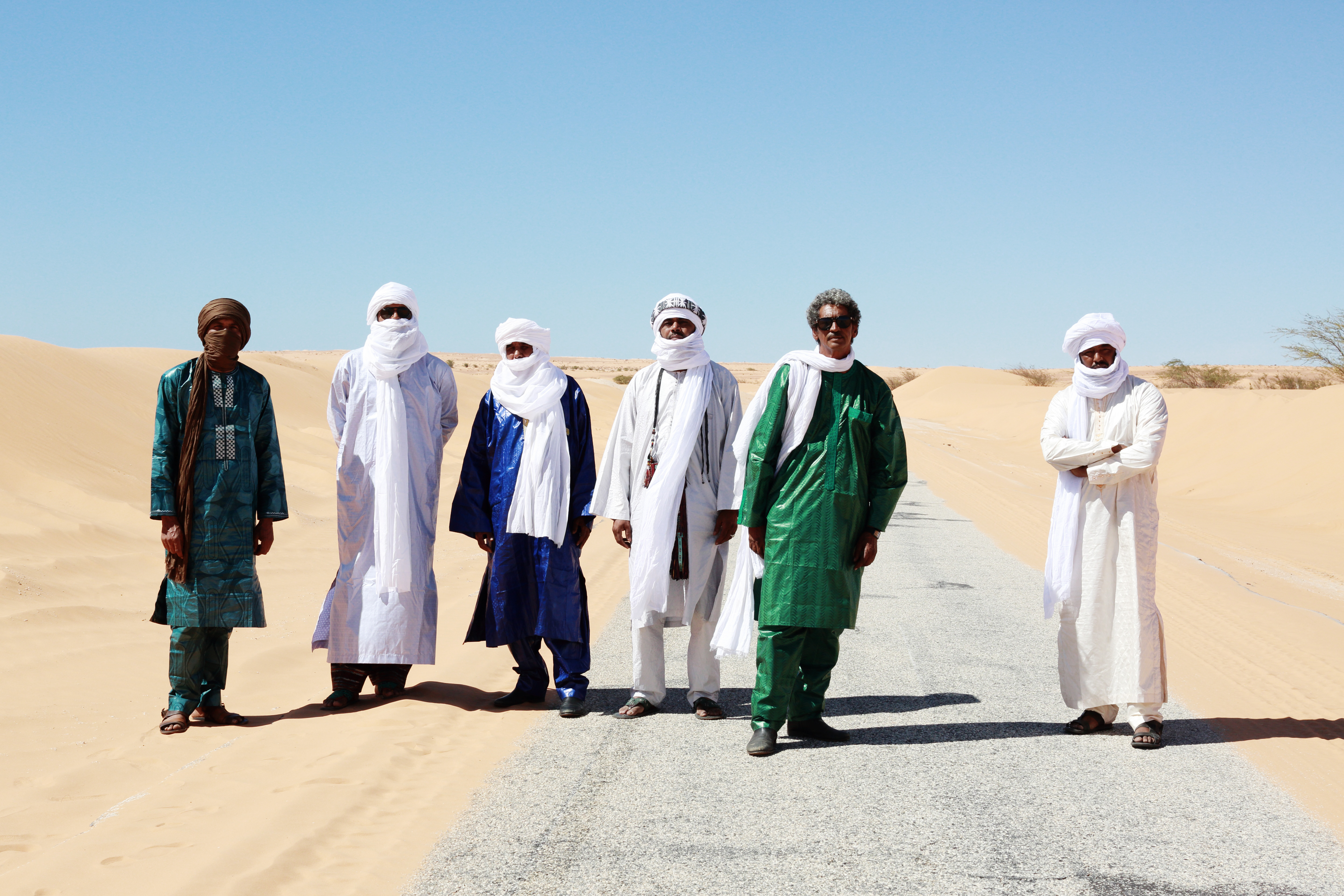 Six individuals, standing in the desert and wearing shawls, face the camera
