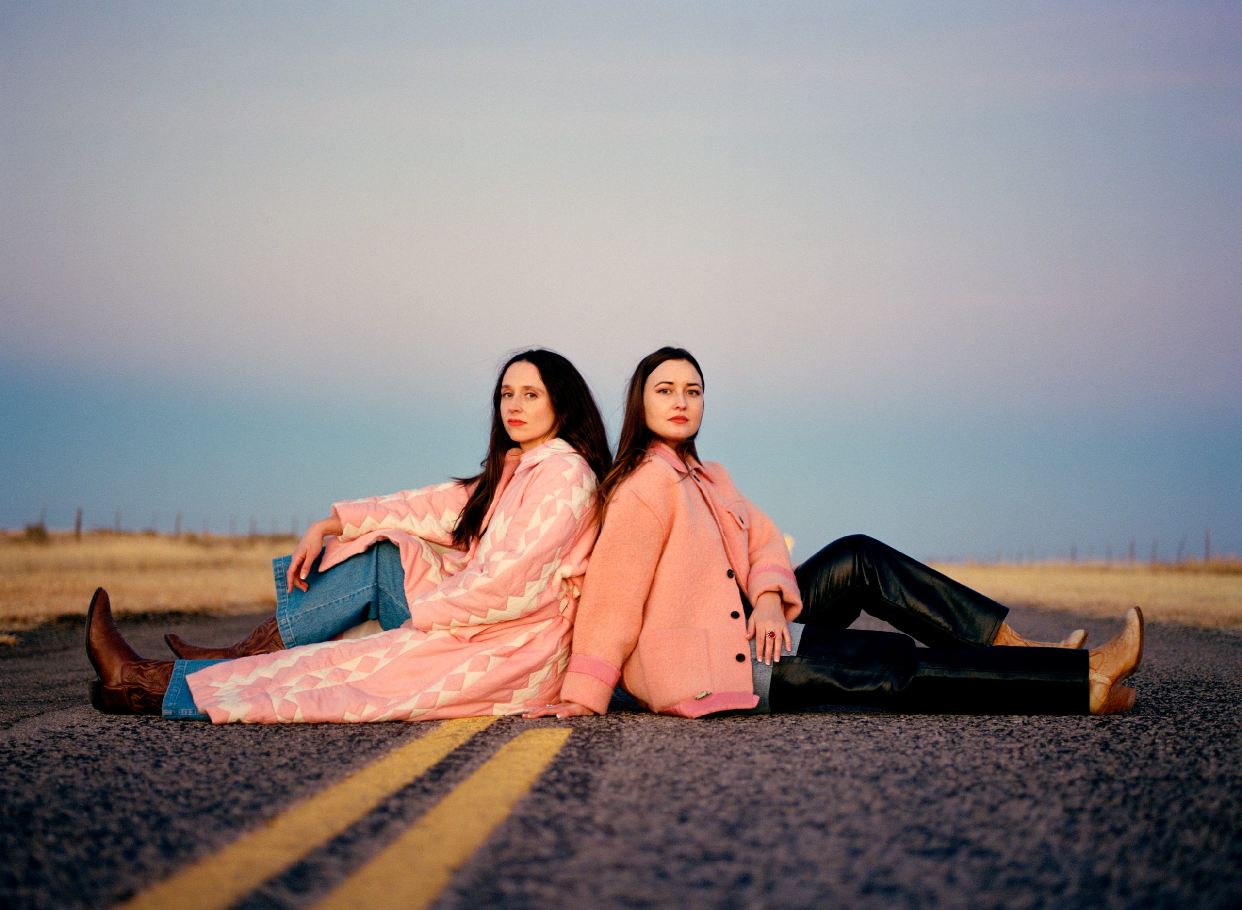 Two women, both wearing pink coats, sit on a road, both facing the camera