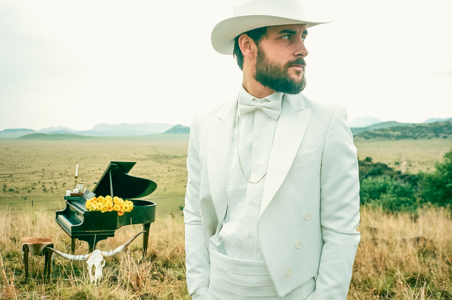 Robert Ellis, standing in a field with a grand piano behind him, looks off to the right