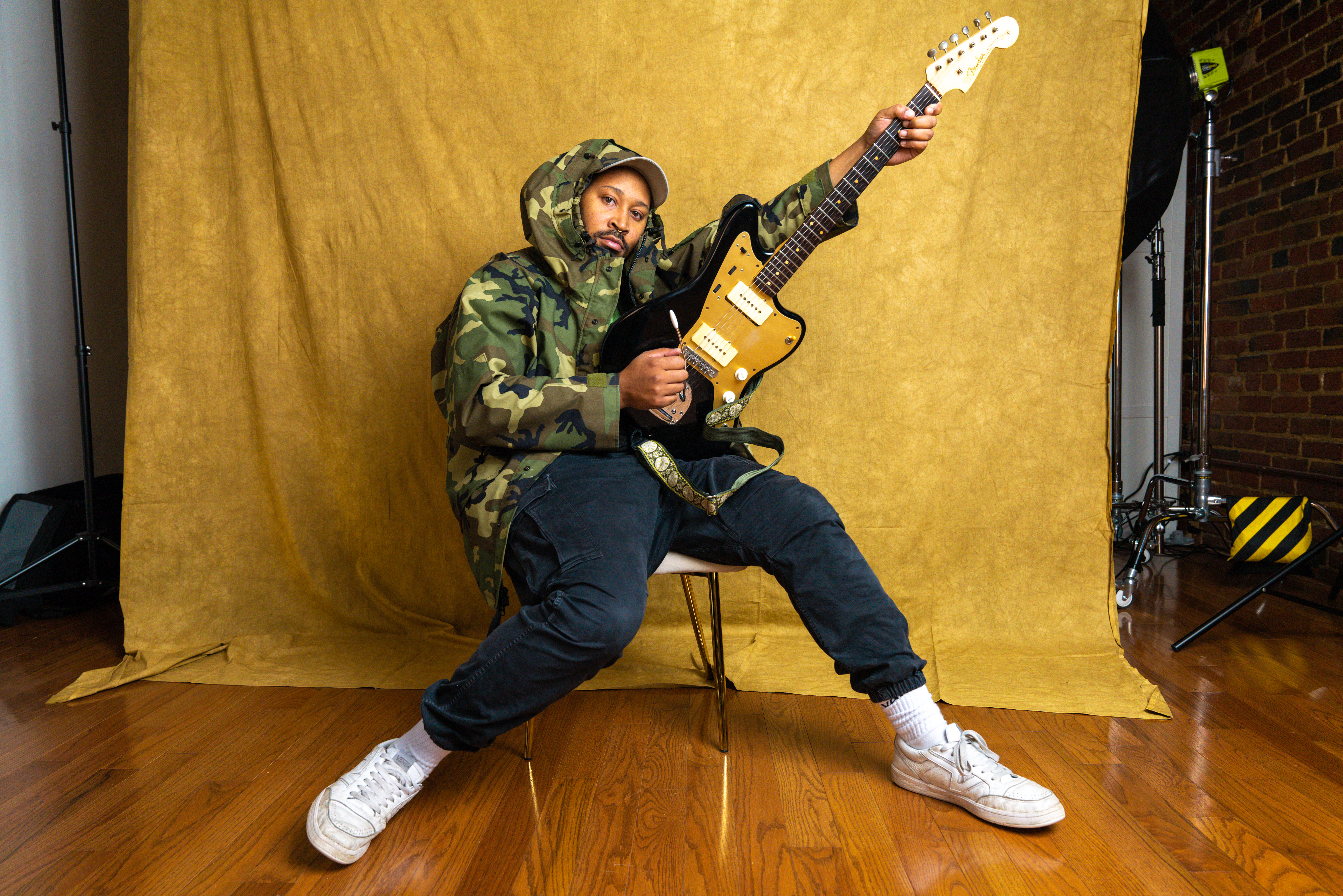 Sitting in front of a yellow backdrop, Bartees Strange holds an electric guitar