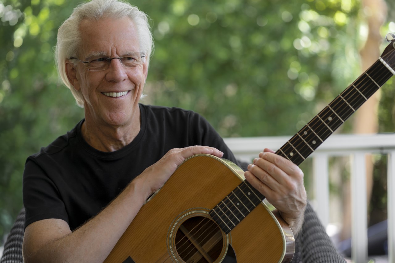Jim Ed Norman, wearing a black t-shirt, holds an acoustic guitar and smiles