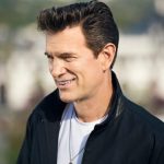 Chris Isaak, wearing a black jacket and white shirt, looks off to the left