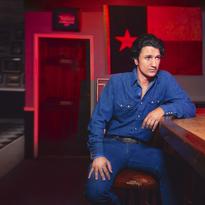 Wearing a denim shirt and jeans, Drake Milligan is seated at a bar, looking off to the right