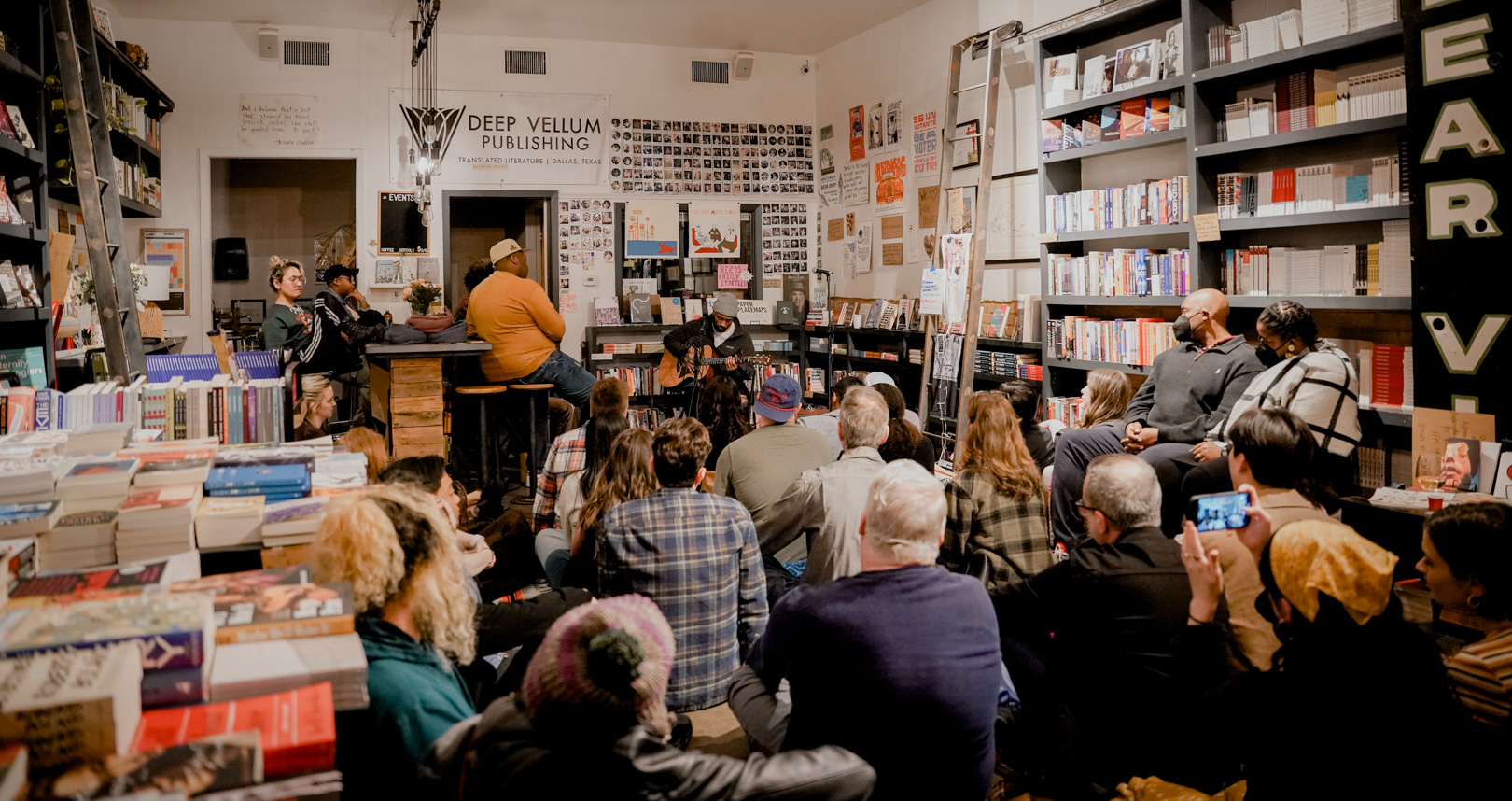 A full bookstore with a person sitting and playing guitar to a large audience
