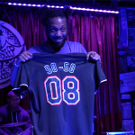 A man holding up a jersey that says "SO-SO" with the number "08"