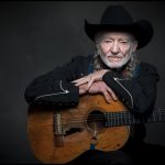 Willie Nelson, dressed in black, faces the camera with Trigger in his lap