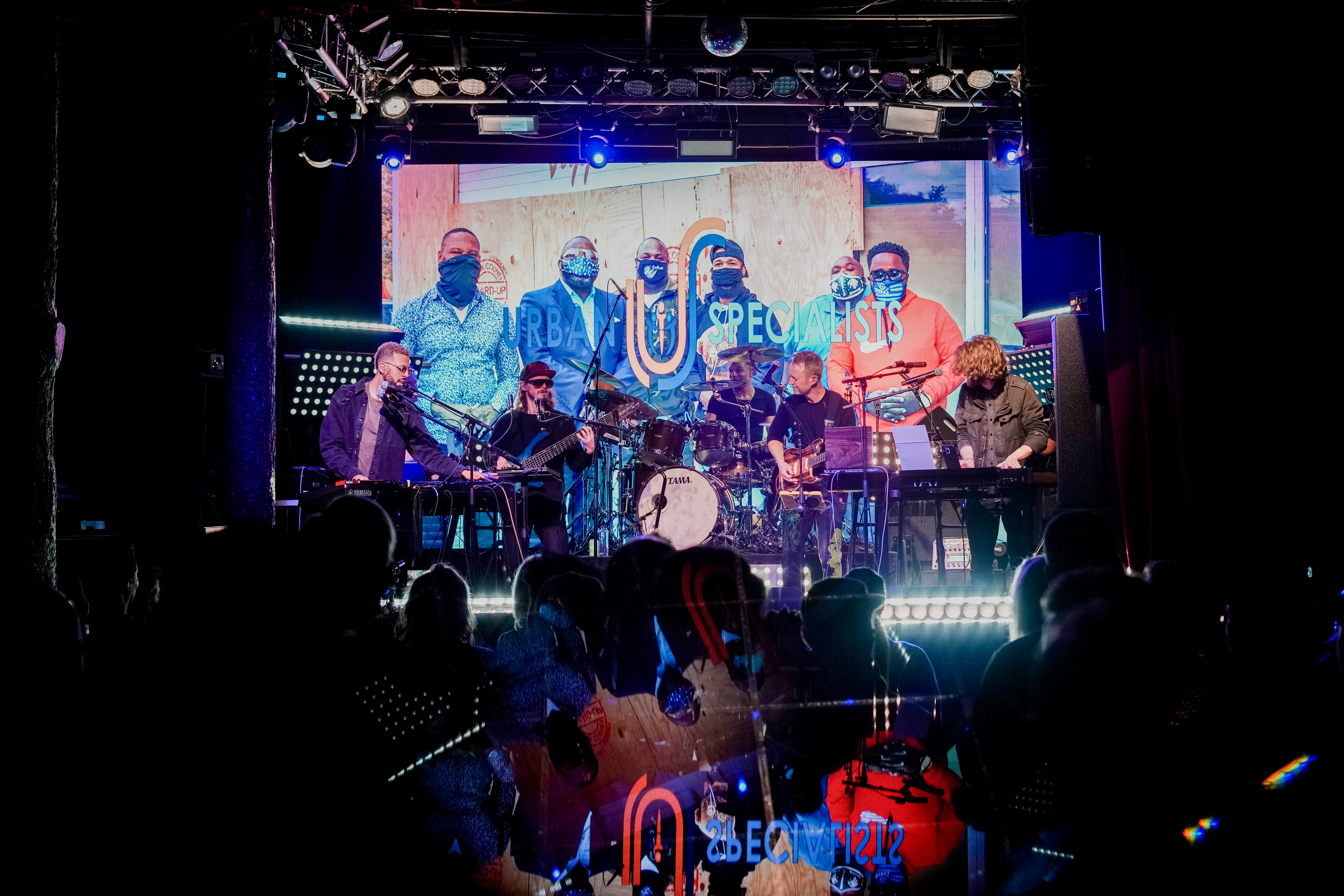 A full band on stage with a projection screen showing a photo of a group of people