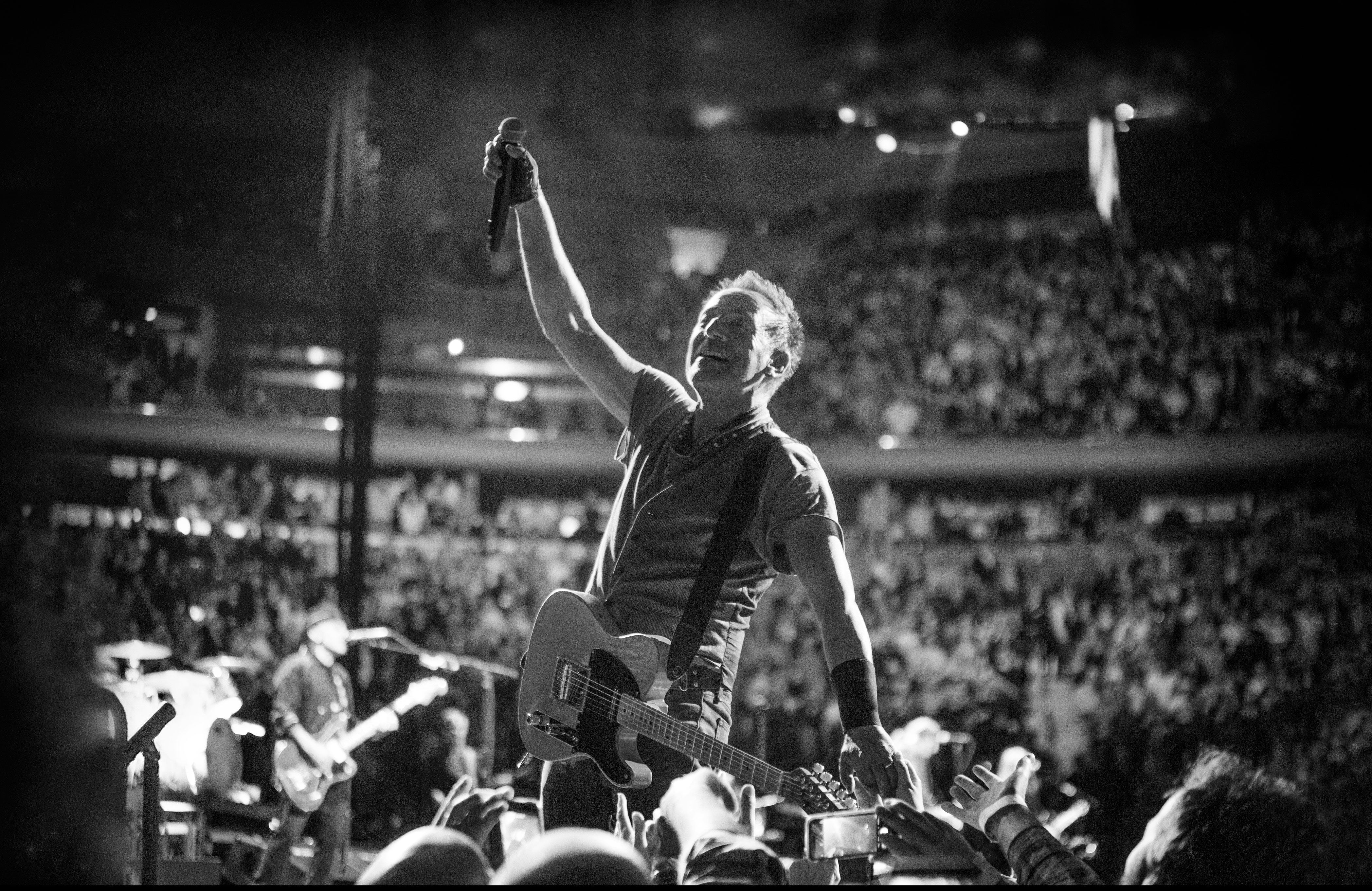 Bruce Springsteen, his electric guitar slung around him, raises a microphone in the air