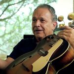 Wearing a black shirt, Jimmie Vaughan holds an acoustic guitar