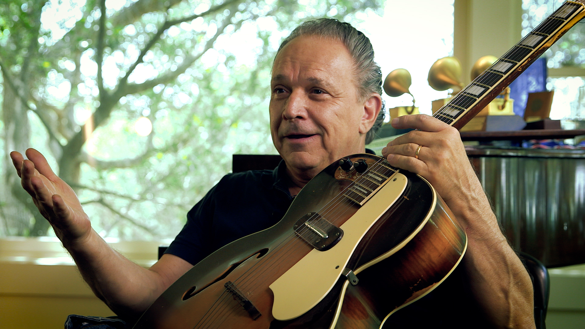 Wearing a black shirt, Jimmie Vaughan holds an acoustic guitar