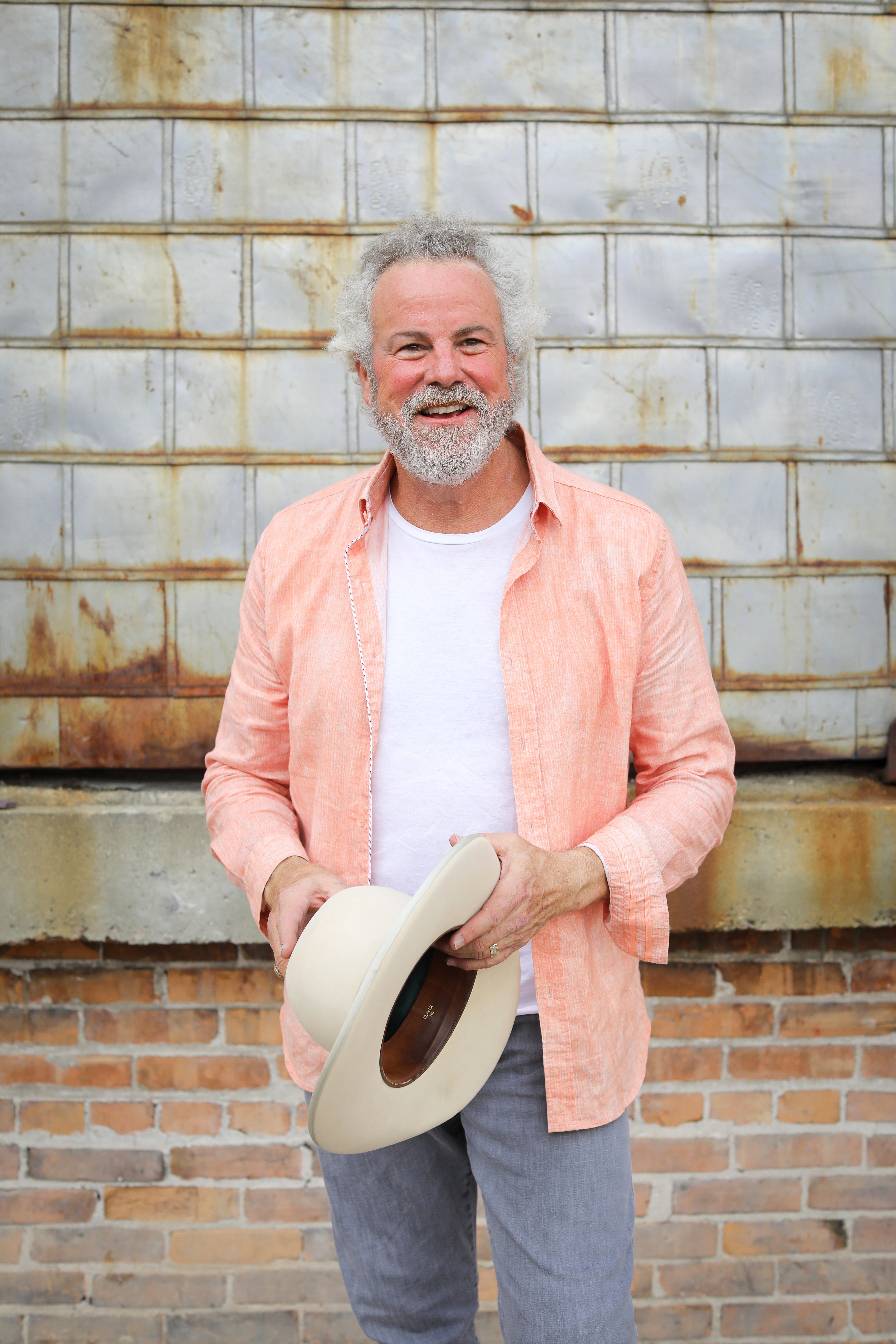 Wearing a peach-colored shirt and holding a hat, Robert Earl Keen smiles for the camera