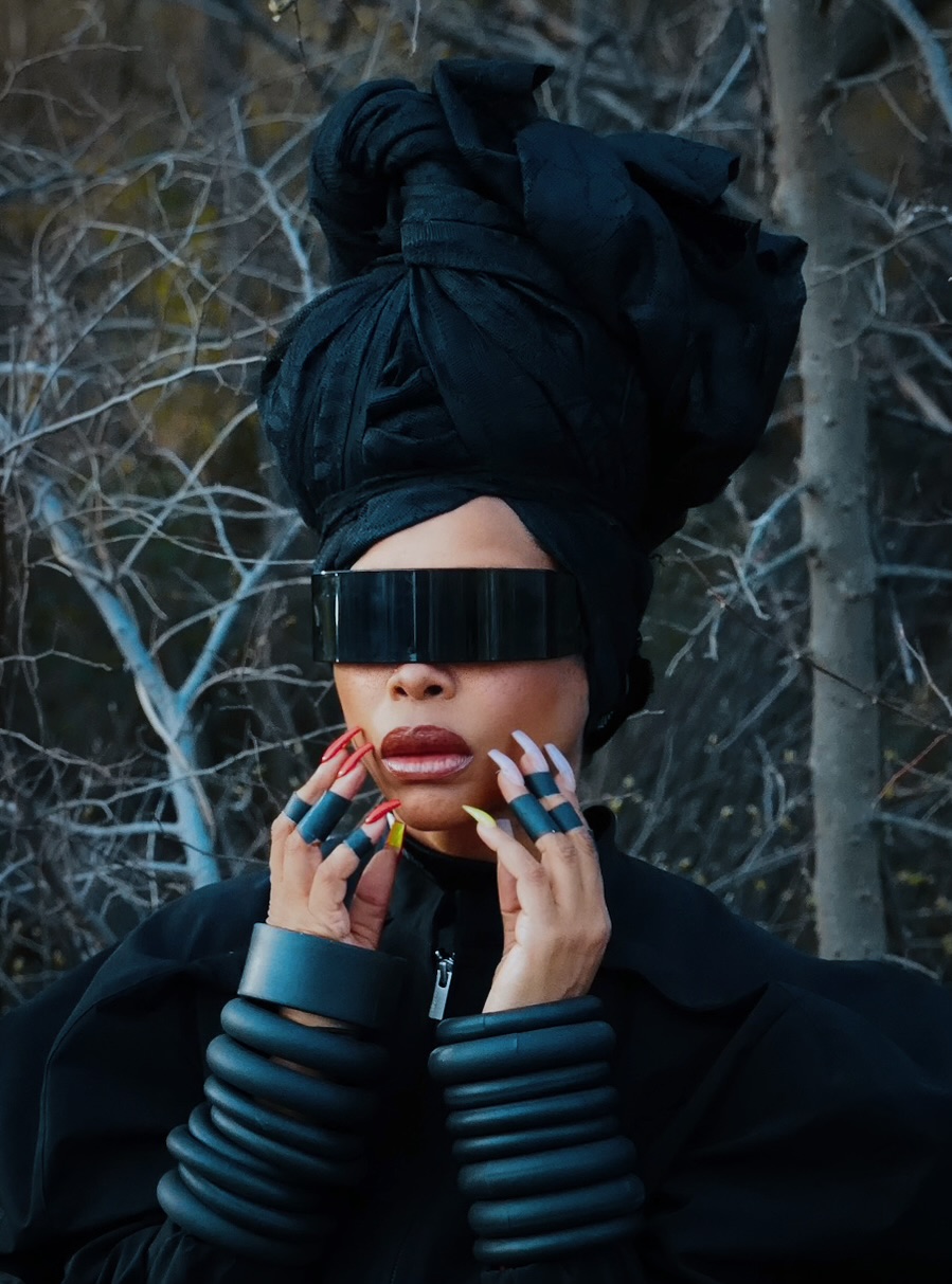 Erykah Badu, wearing a black head wrap and black glasses, holds her hands to her face