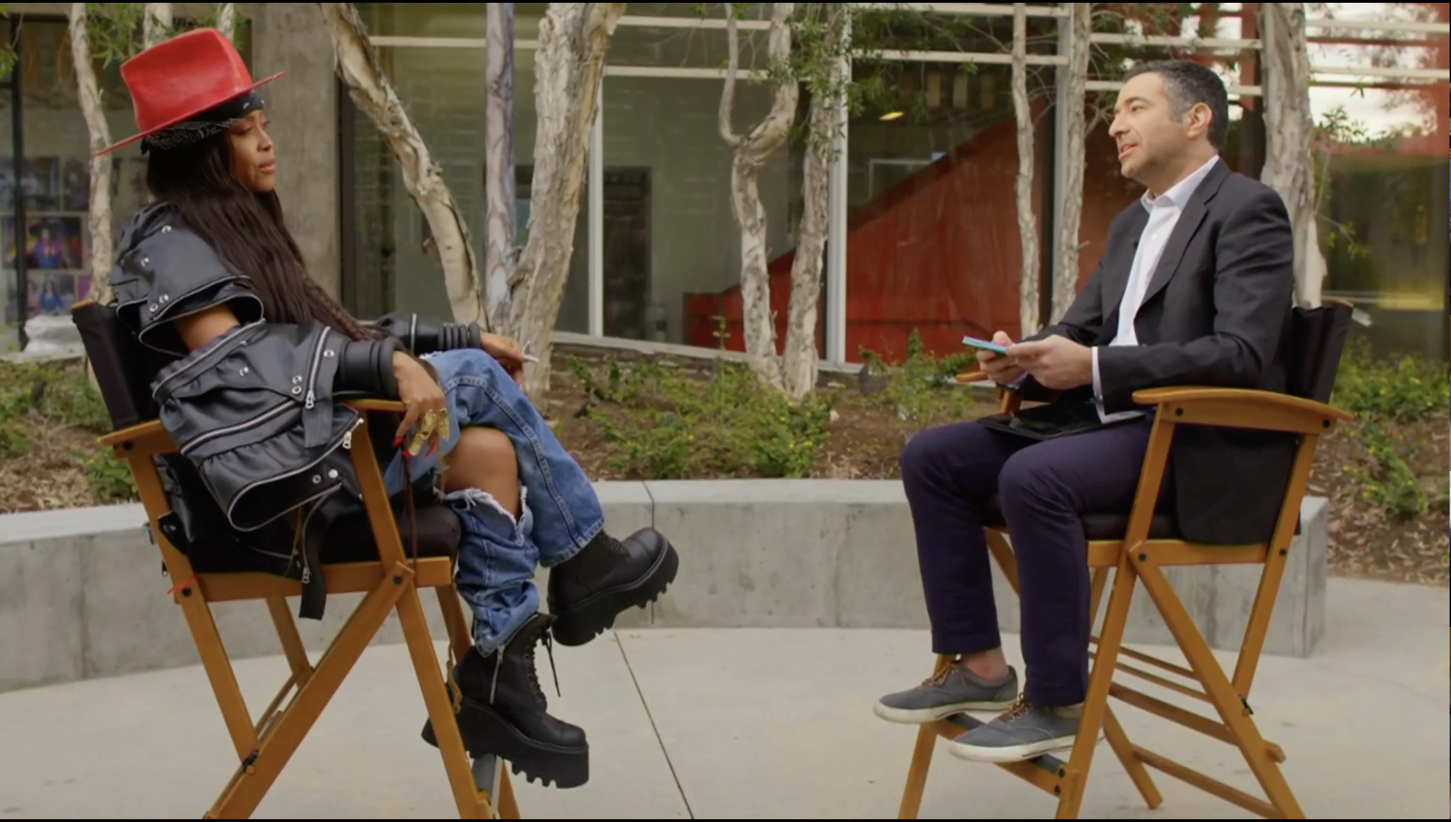 Erykah Badu and Ari Melber face each other in chairs on an outdoor patio.