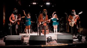 A full band on stage wearing gorilla outfits, and two dancers