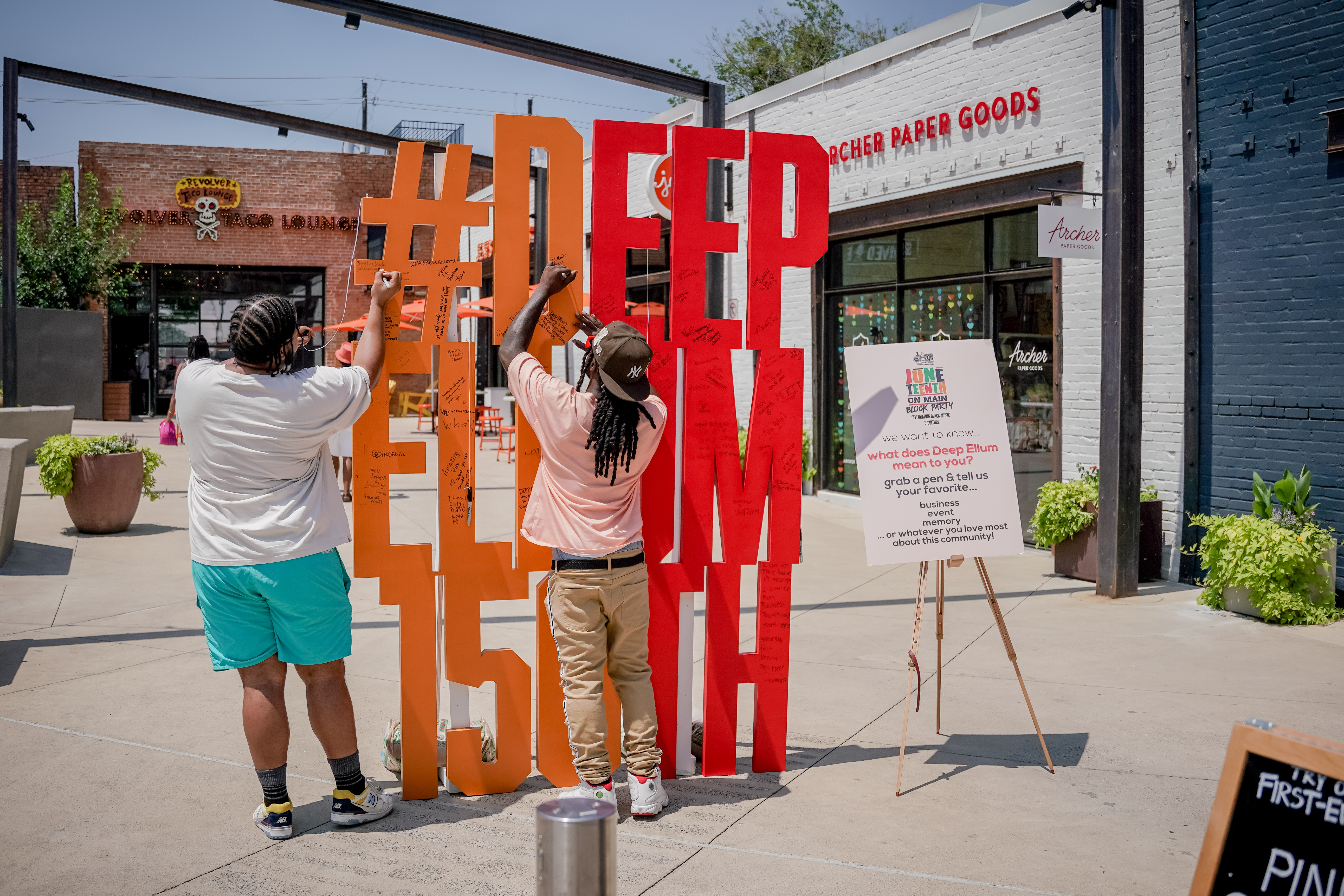 A painted wooden structure that says "#DeepEllum150th," people writing with sharpies on it