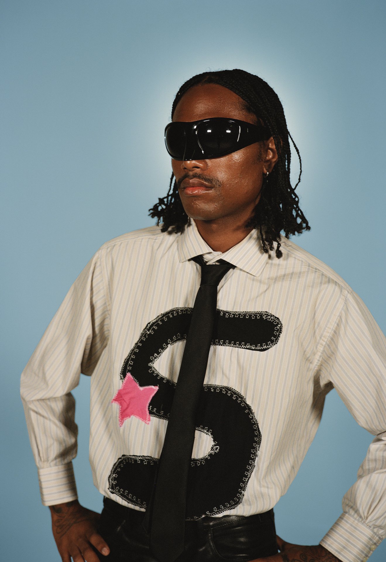 Steve Lacy, wearing dark glasses and a shirt with a giant dollar sign, faces the camera against a blue wall
