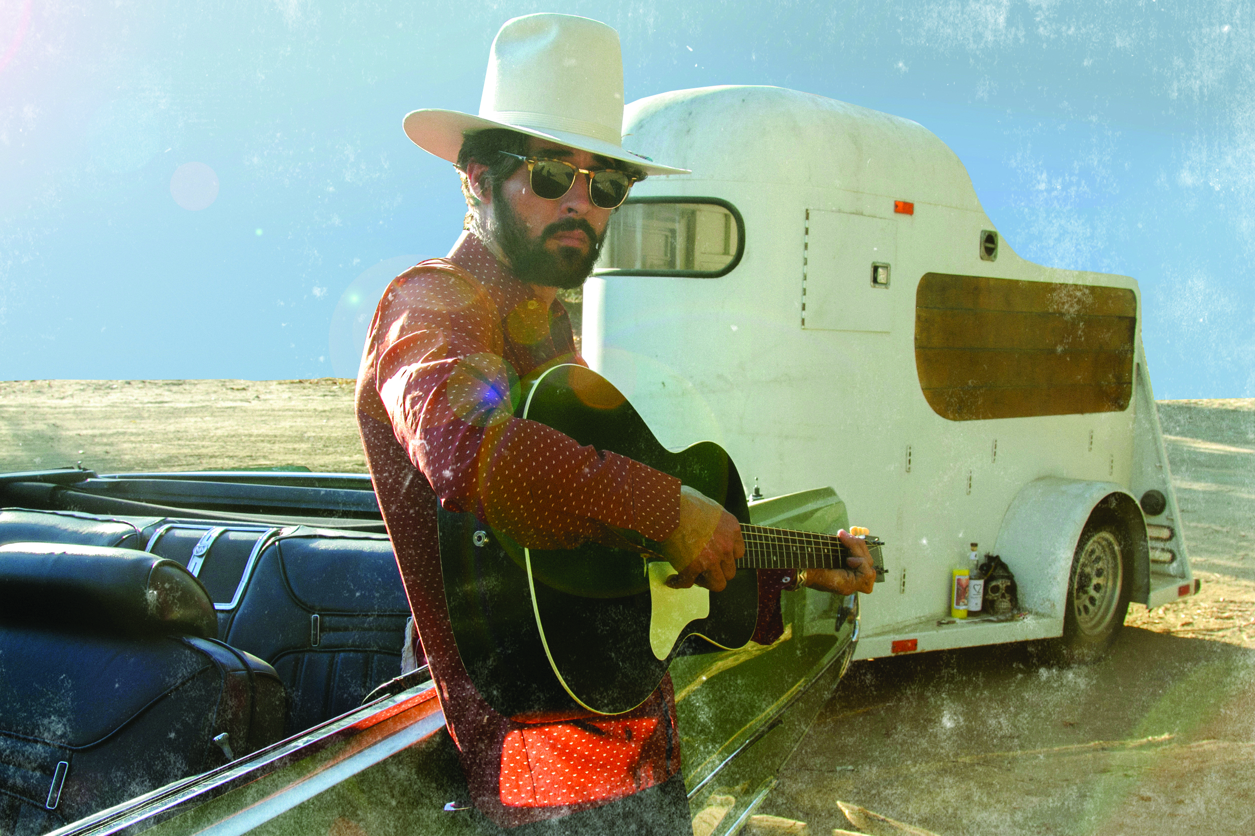 Holding a guitar and wearing sunglasses, Ryan Bingham stands in front of an old truck