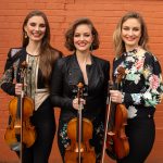 Holding their fiddles, the three Quebe sisters stand in front of an orange wall