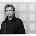 Andy Summers, wearing a black jacket, stands against a wall with a grid pattern on it