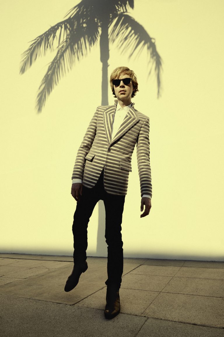 Wearing sunglasses and a striped suit, Beck stands in front of a wall with the shadow of a palm tree on it
