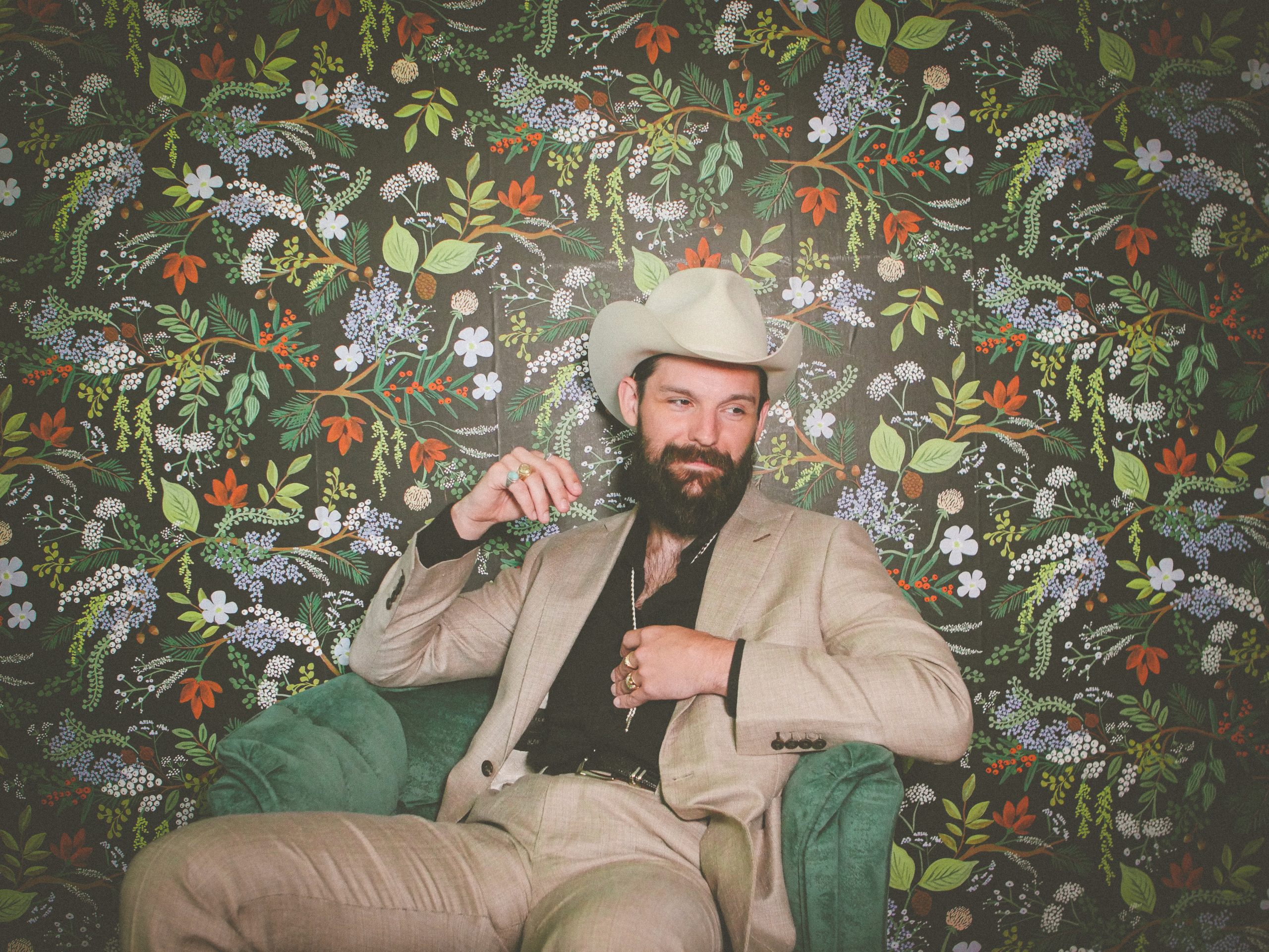Wearing a tan suit, Nathan Mongol Wells sits in front of tropical wallpaper