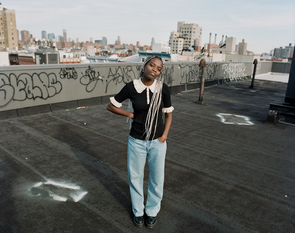 Hannah Jadagu, wearing a black top and blue jeans, stands on a NYC rooftop