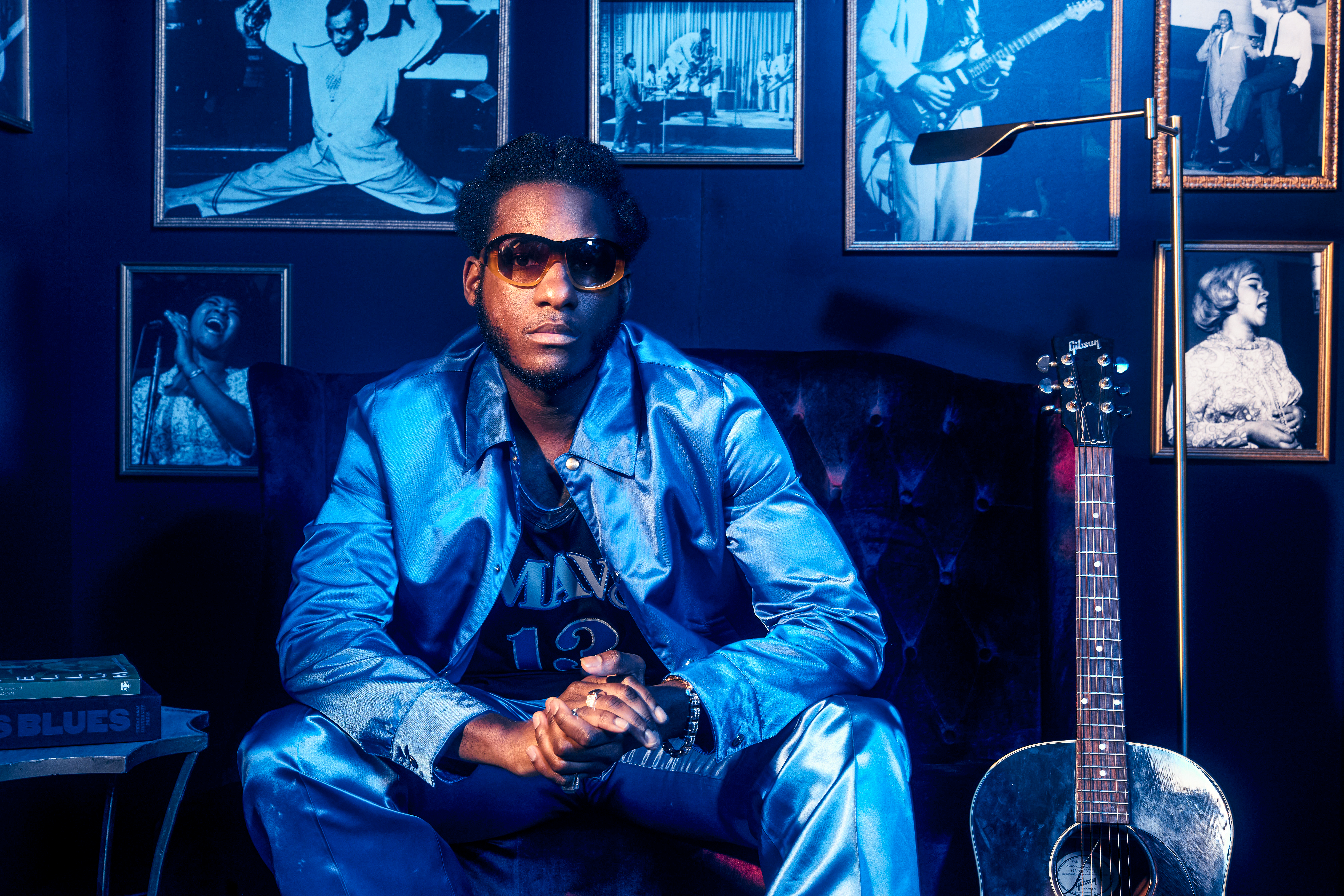 Leon Bridges, wearing a denim jacket and a basketball jersey, sits on a couch