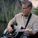 Don Henley, holding an acoustic guitar, sits amid a swamp