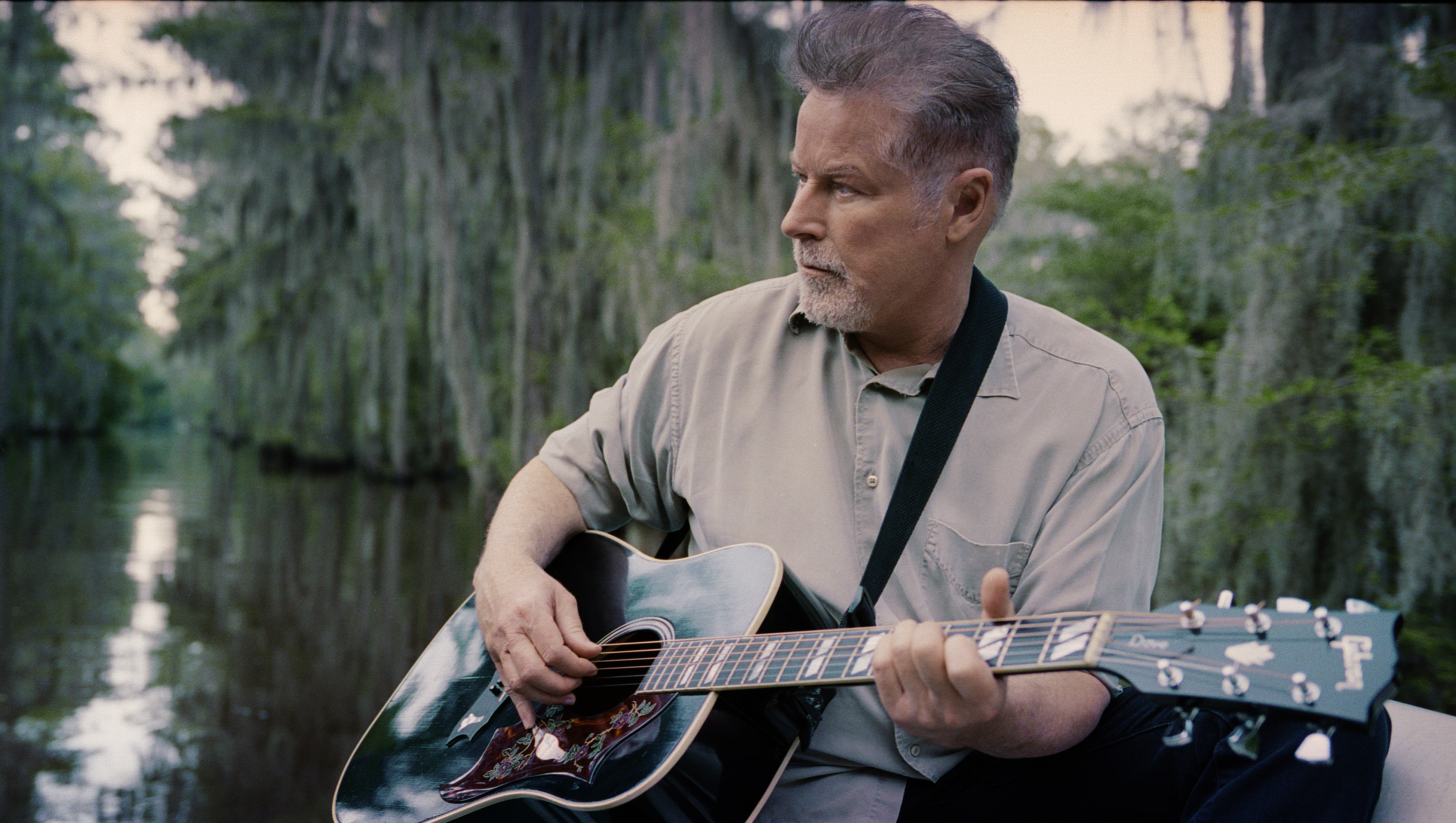 Don Henley, holding an acoustic guitar, sits amid a swamp