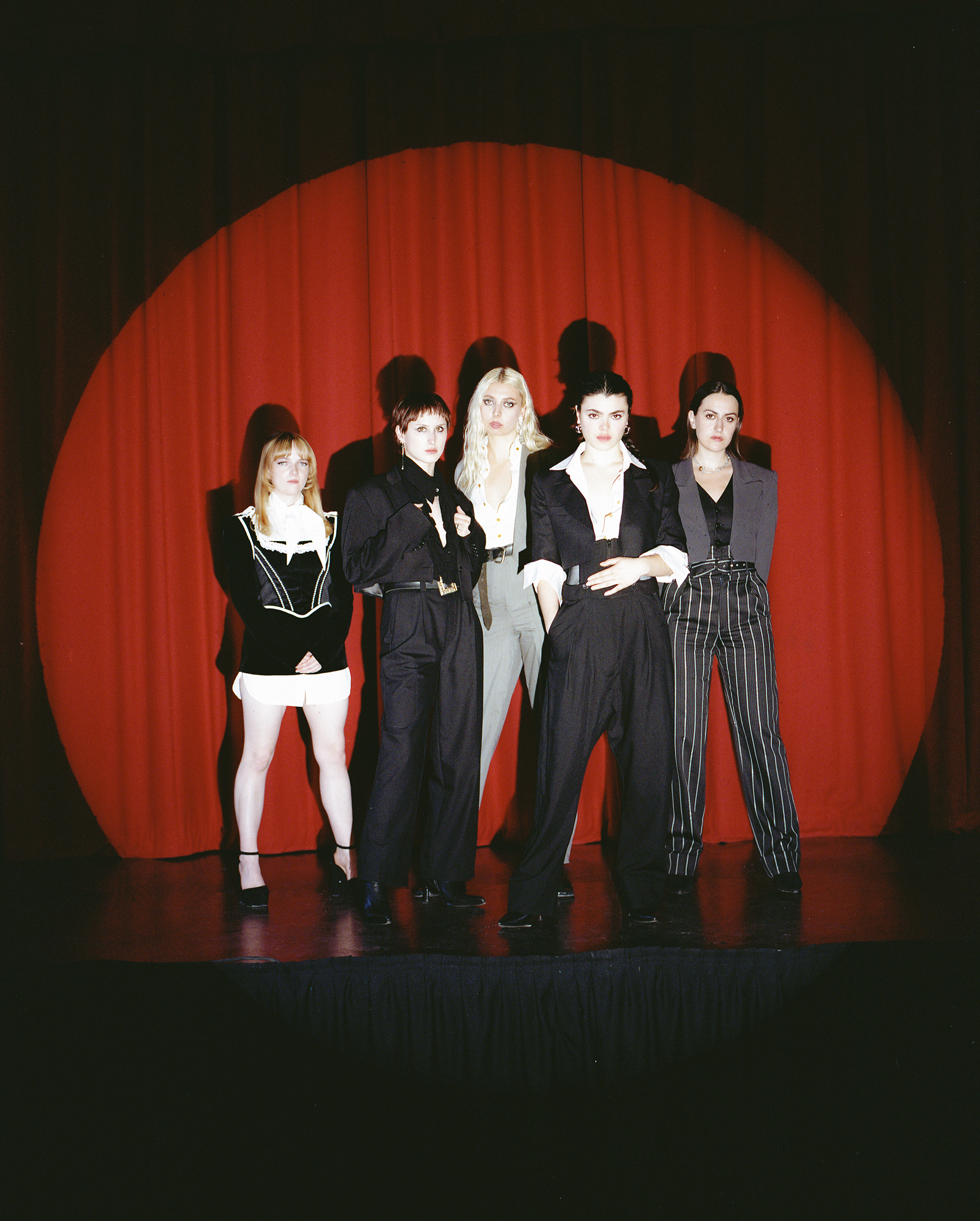 The five members of the Last Dinner Party stand in a spotlighted circle, with a red curtain behind them