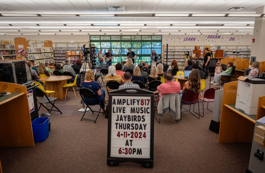 A sign that says "Amplify Live Music Jaybirds" in front of a seated crowd inside a library