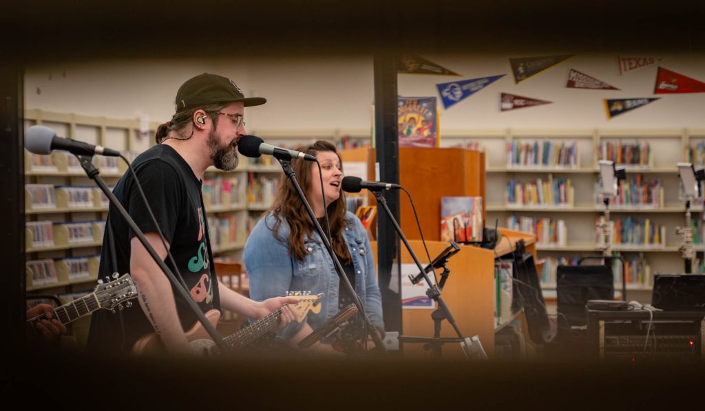Two musicians singing in a library, as seen through two bookshelves